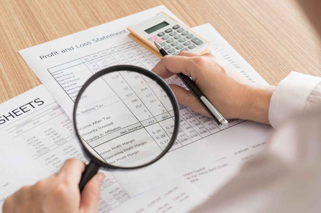 Person holding magnifying glass looking at a profit and loss statement.